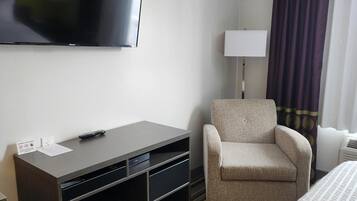 Quarto de hotel com televisão de tela plana na parede, uma mesa com controle remoto, uma cadeira, um abajur e uma cama com carpete listrado.