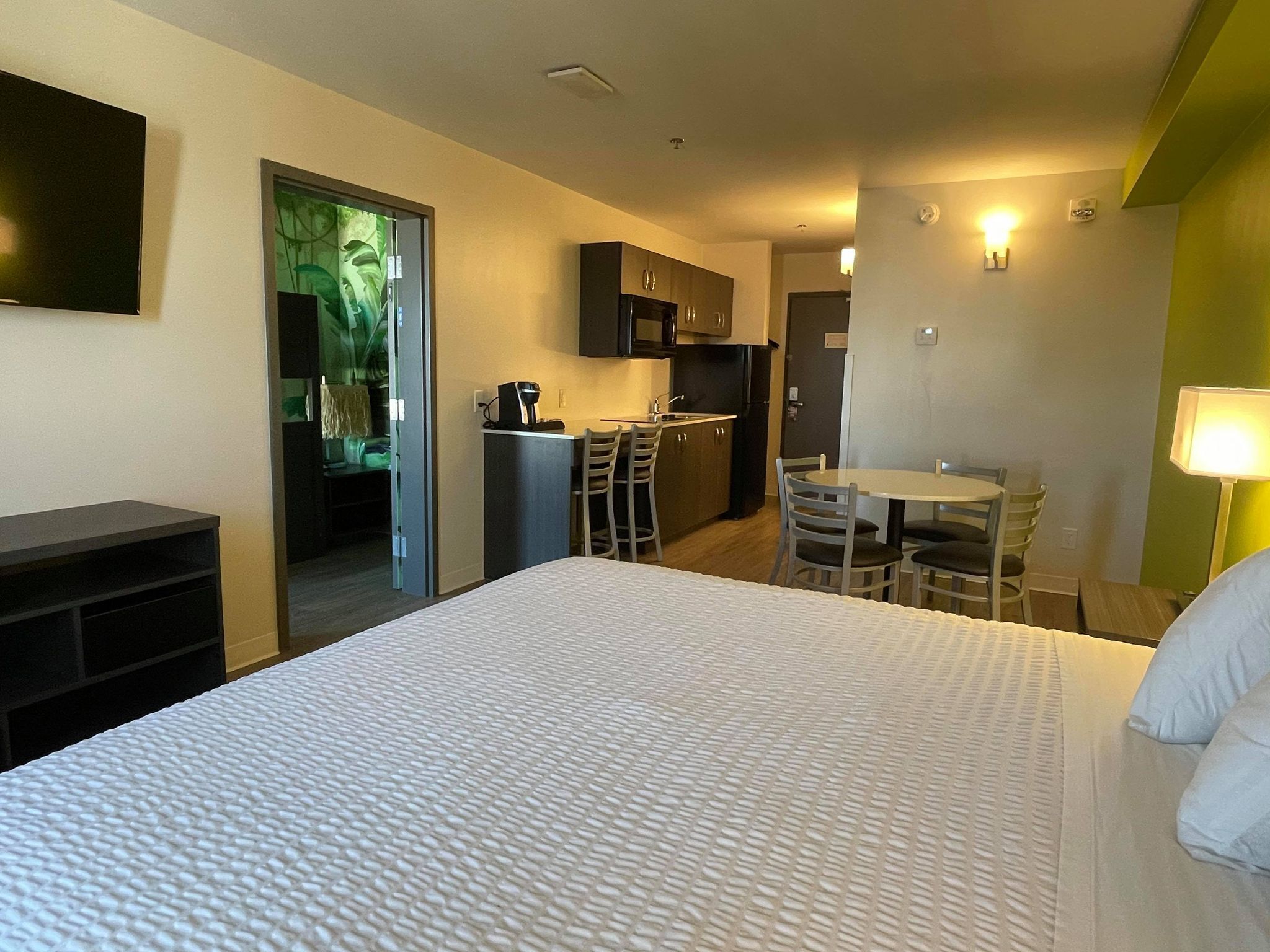 A hotel room with a bed, a desk, a chair, a small kitchenette, and a TV.