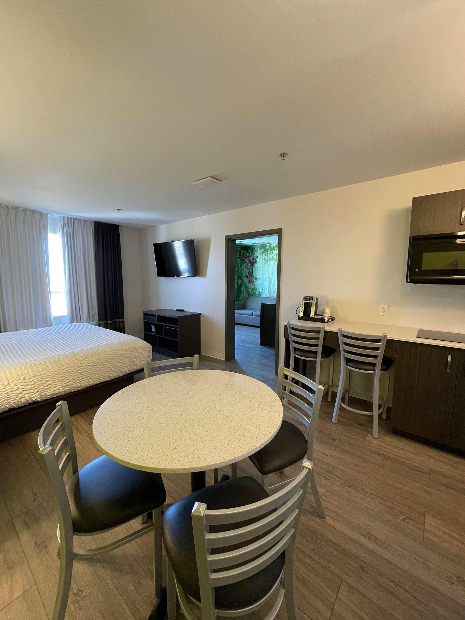 A modern hotel room with a bed, a round dining table, and a kitchenette.