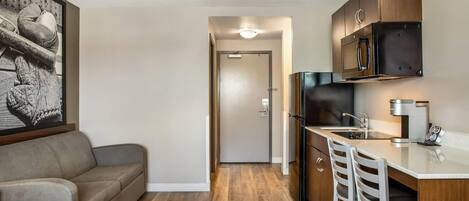 A compact kitchen with a refrigerator, microwave, and coffee maker, a kitchenette with a sink and bar stools, a sofa, and a framed picture on the wall.