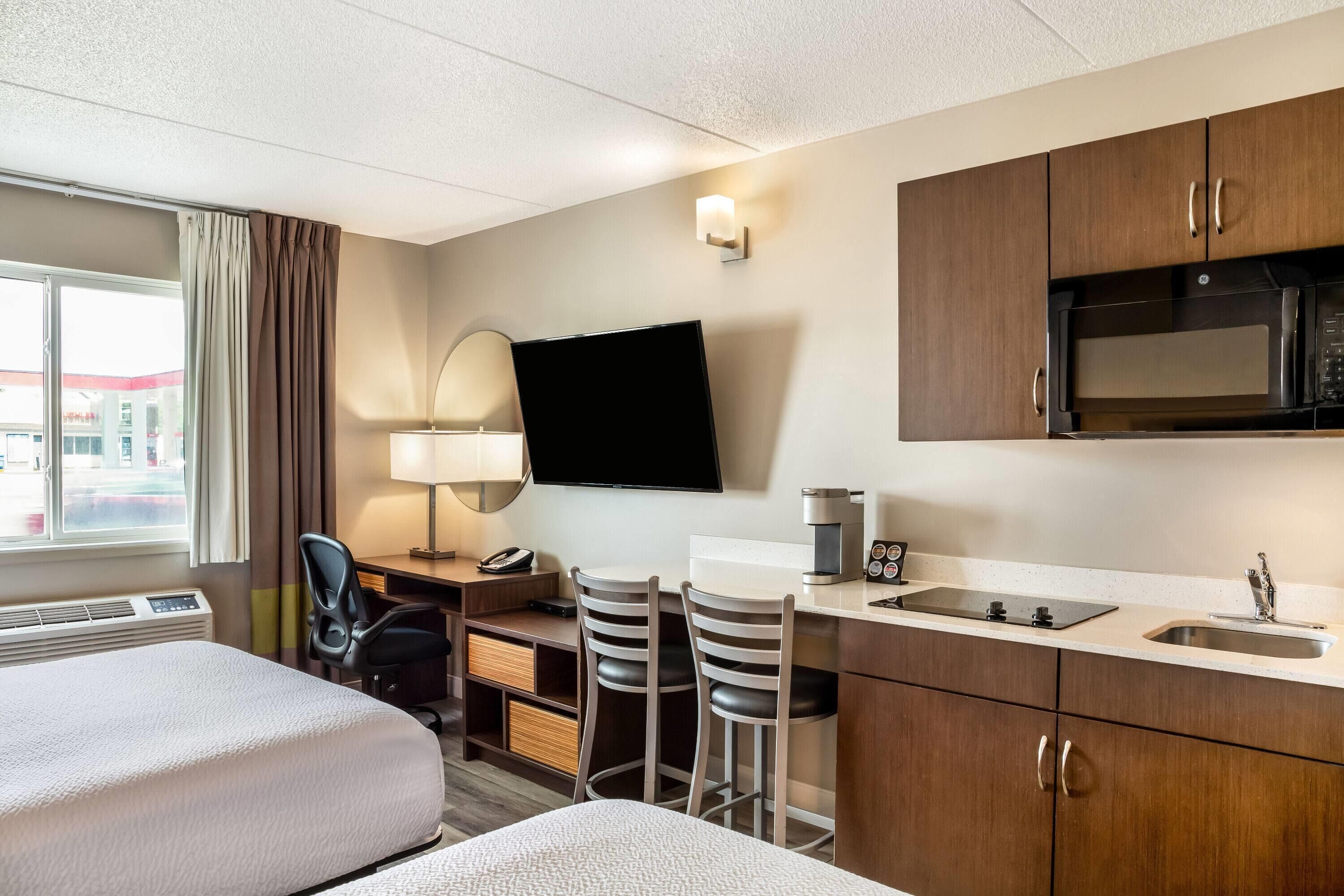 A hotel room with a bed, desk, chairs, a TV, a microwave, and a sink.