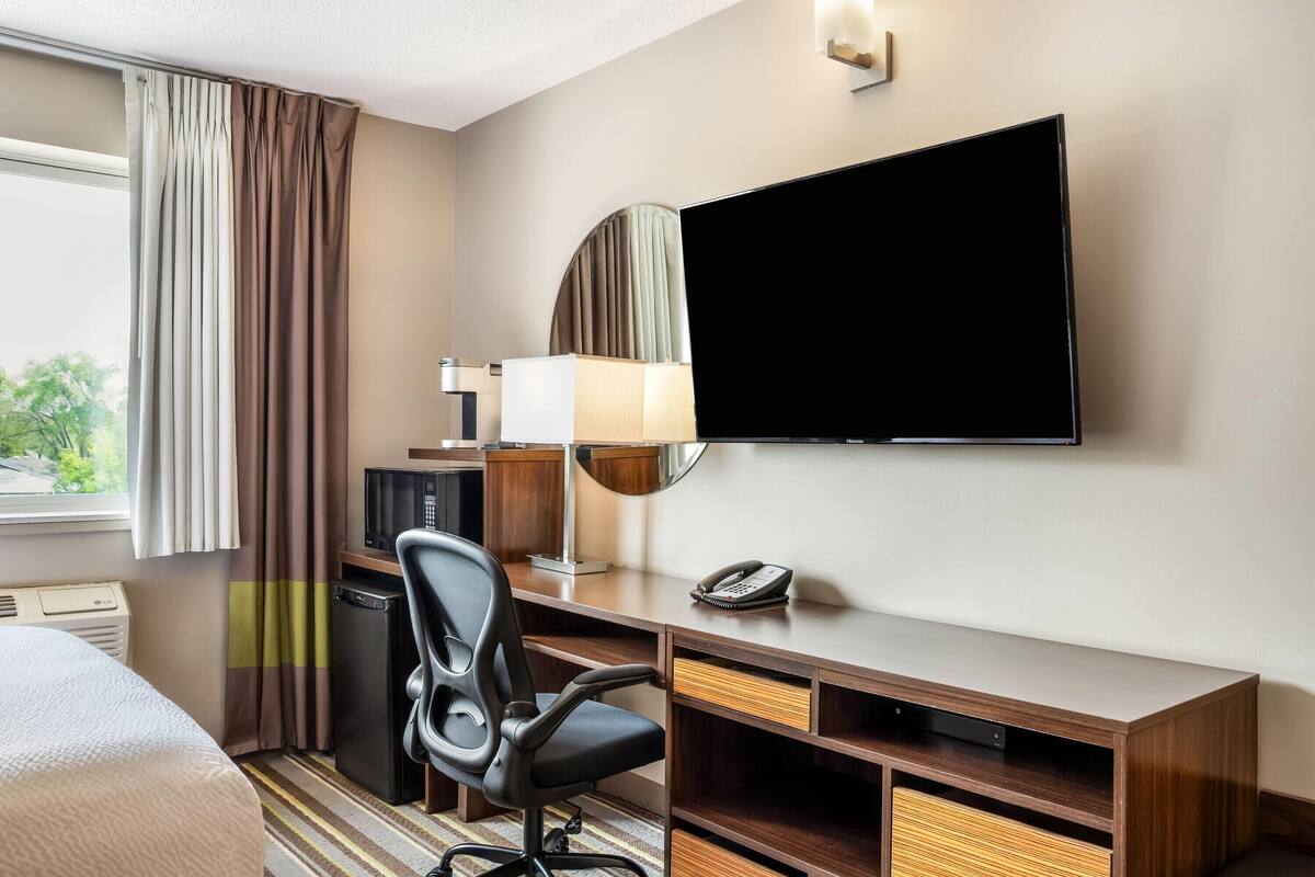 Ein Hotelzimmer mit Bett, Schreibtisch, Stuhl, Fernseher, Lampe, Telefon und Blick auf Bäume.