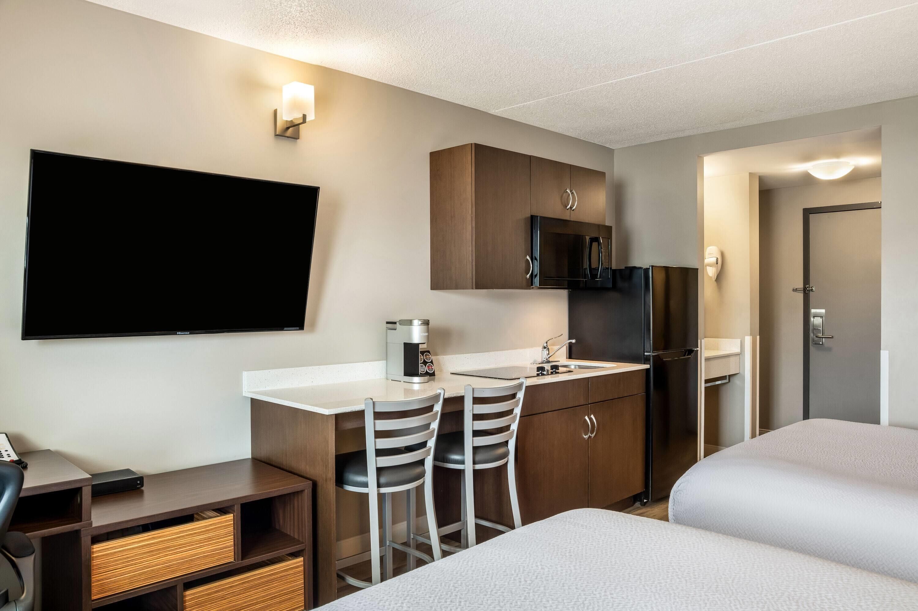 A hotel room with a bed, a desk with two chairs, a TV, a microwave, and a small kitchenette.