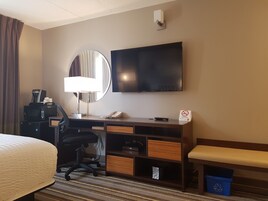 Habitación de hotel con cama, escritorio con lámpara, televisor, cafetera, teléfono, cubo de basura y contenedor de reciclaje.