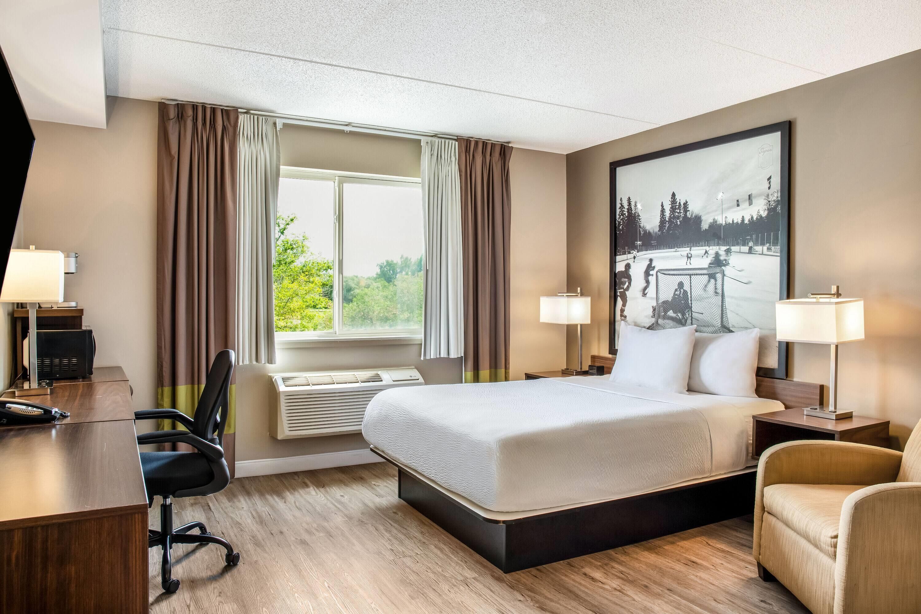 Habitación de hotel con cama, escritorio, silla, aire acondicionado y un amplio ventanal con cortinas.