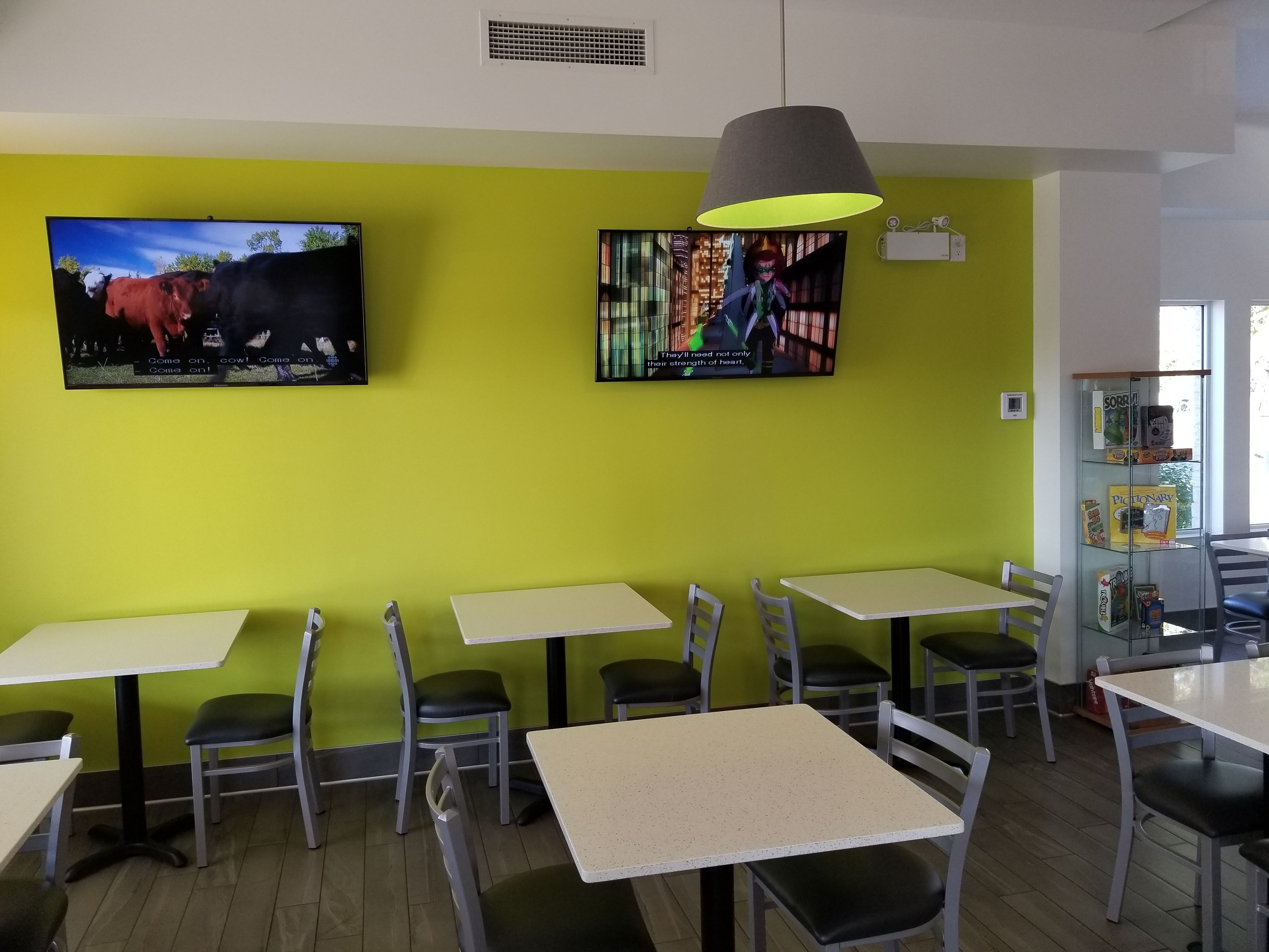 Ein Restaurant mit grünen Wänden, mehreren Tischen und Stühlen sowie zwei Fernsehern, die unterschiedliche Inhalte zeigen.
