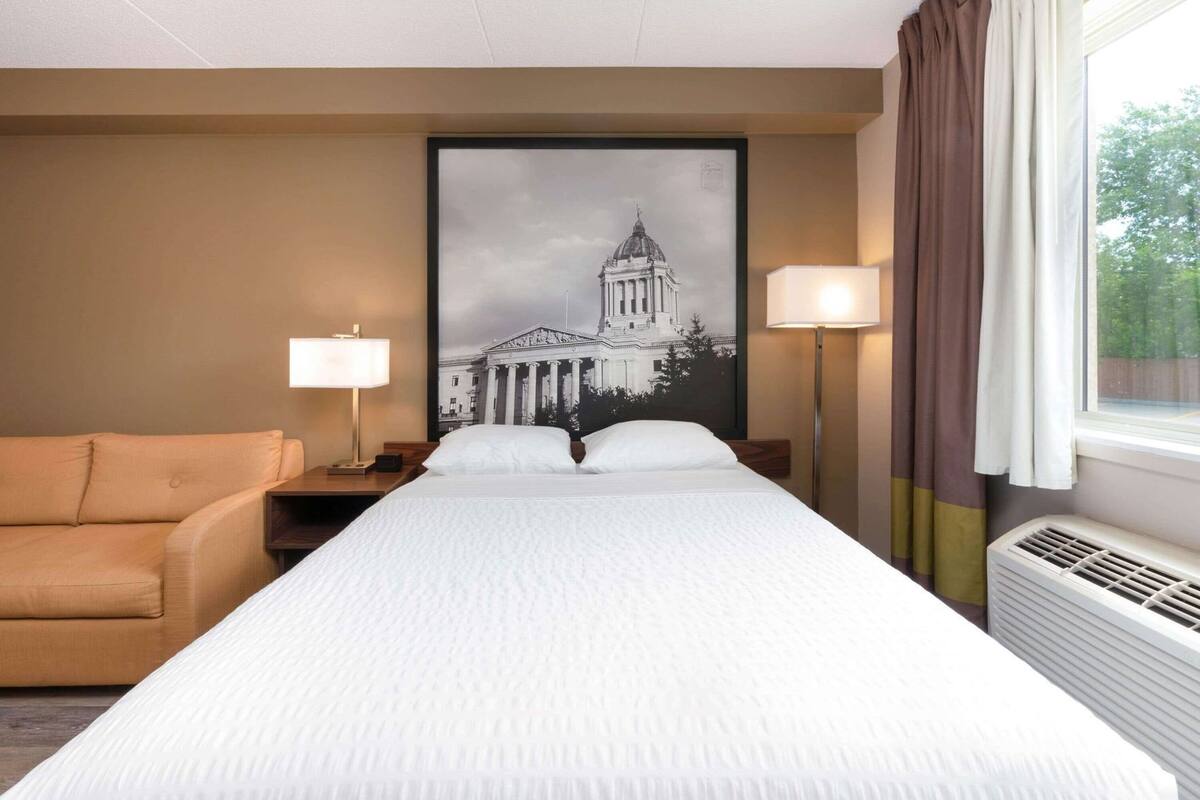 Ein Hotelzimmer mit einem Bett, einem Sofa, zwei Nachttischlampen, einem großen gerahmten Bild eines Gebäudes und einem Fenster mit Vorhängen.