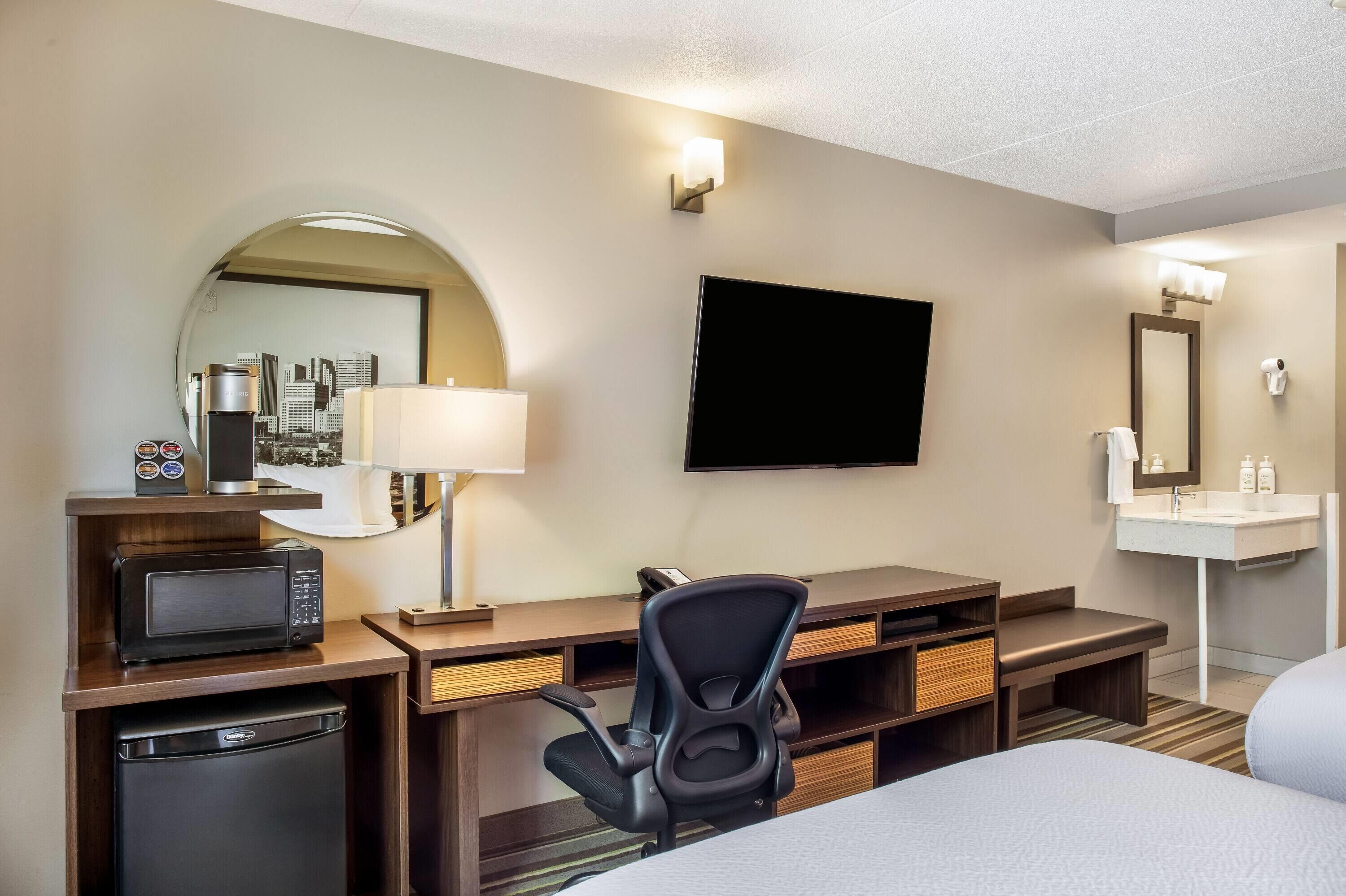 Habitación de hotel con cama, escritorio, microondas, lámpara, televisor y un baño pequeño con lavamanos y espejo.