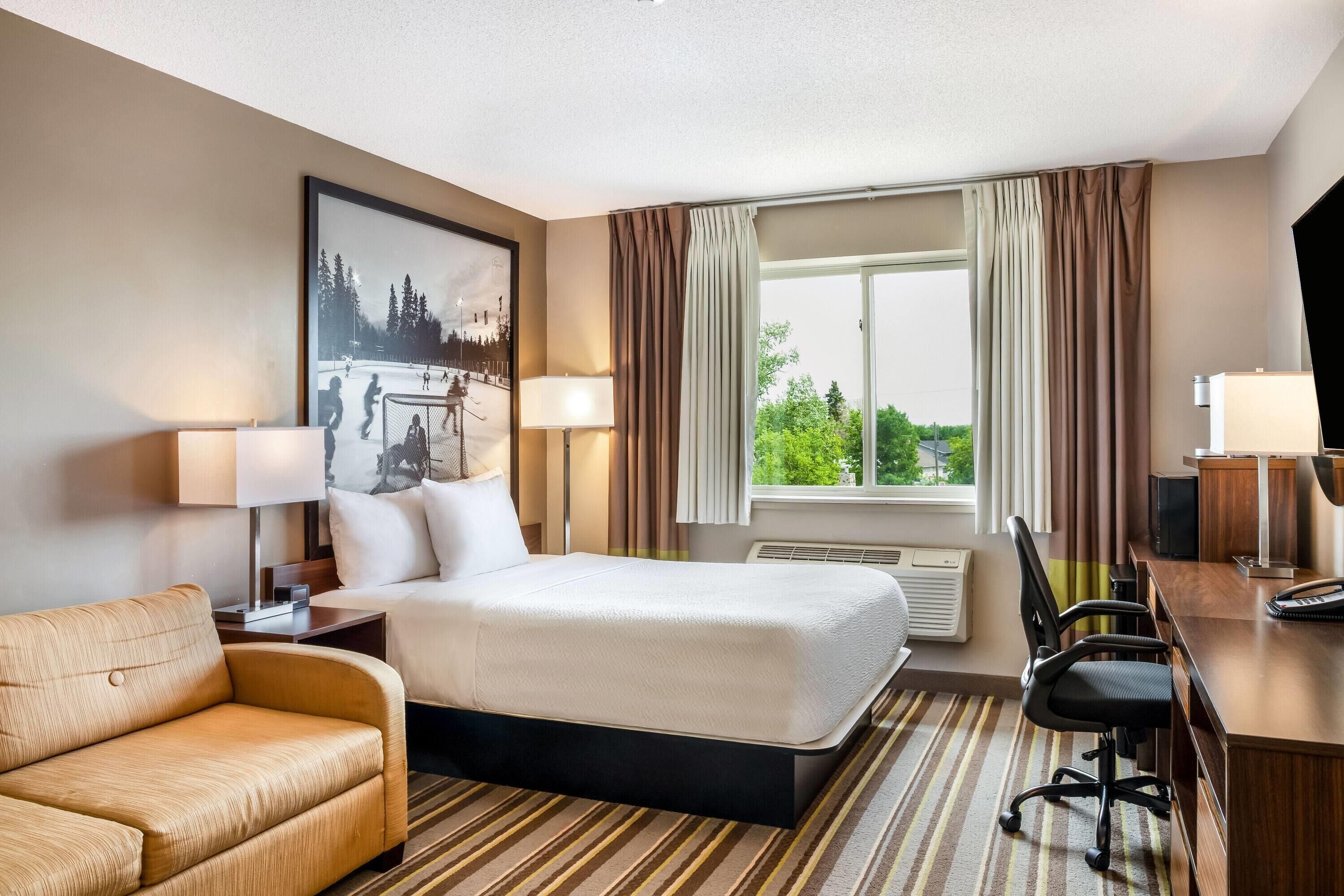 A hotel room with a bed, a desk, a chair, a lamp, a picture on the wall, and a window with curtains.