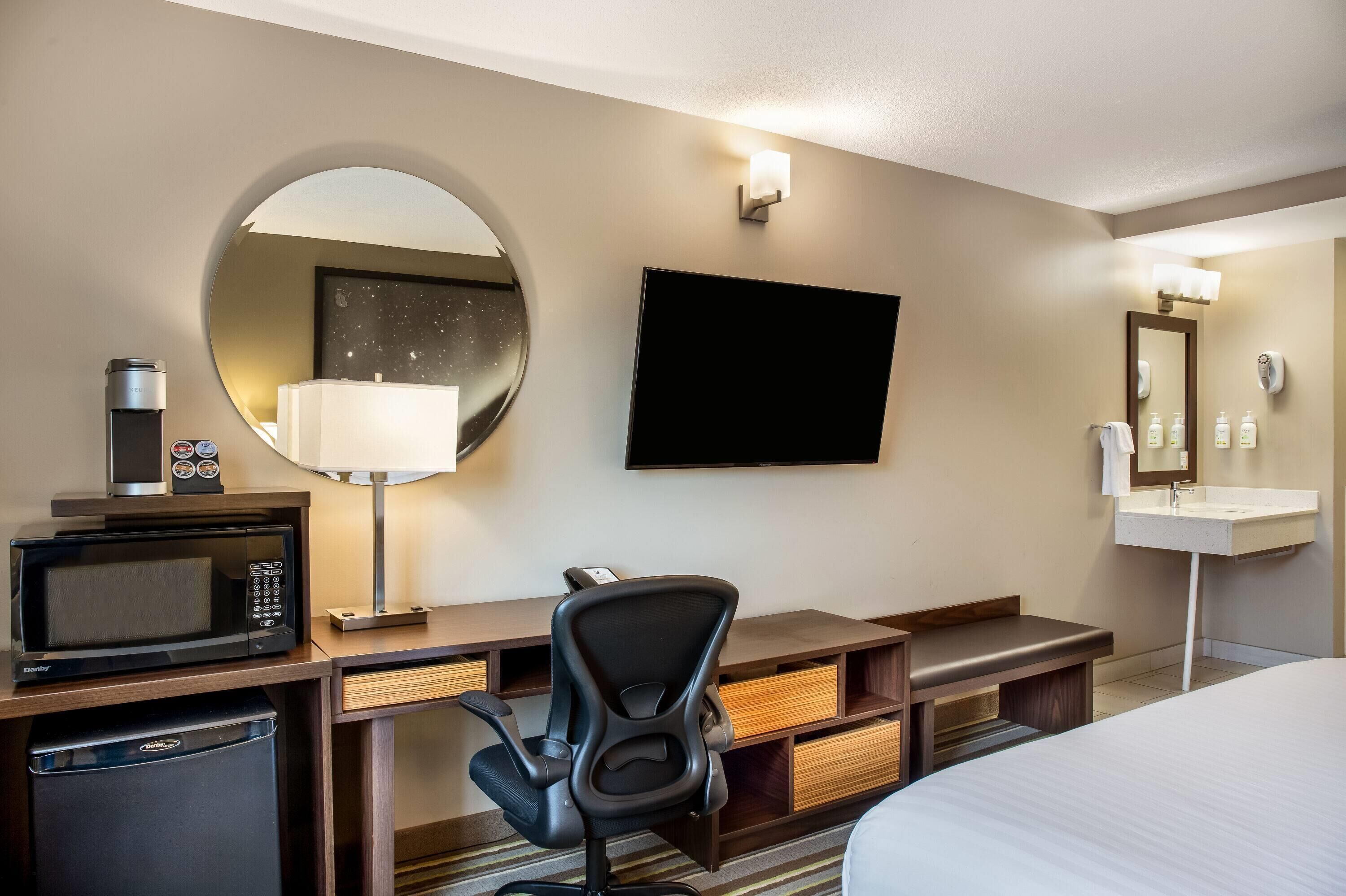 A hotel room with a bed, a desk with a microwave, a chair, a TV, a round mirror, and a sink.