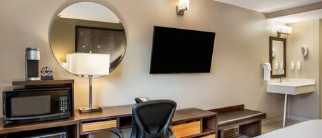 A hotel room with a bed, a desk with a microwave, a chair, a TV, a round mirror, and a sink.