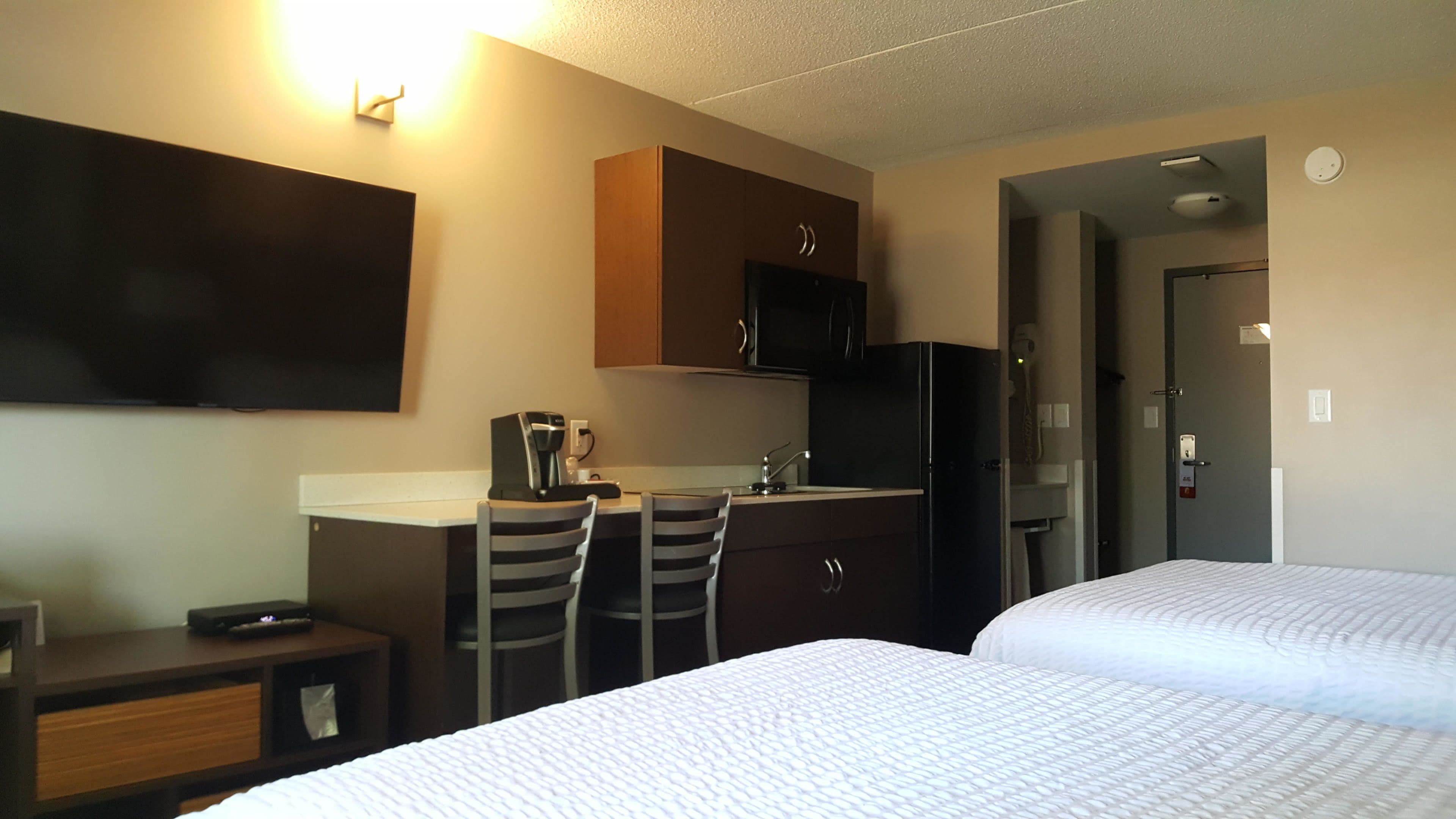 Quarto de hotel com duas camas, uma kitchenette pequena, televisão de tela plana e uma escrivaninha com cadeiras.
