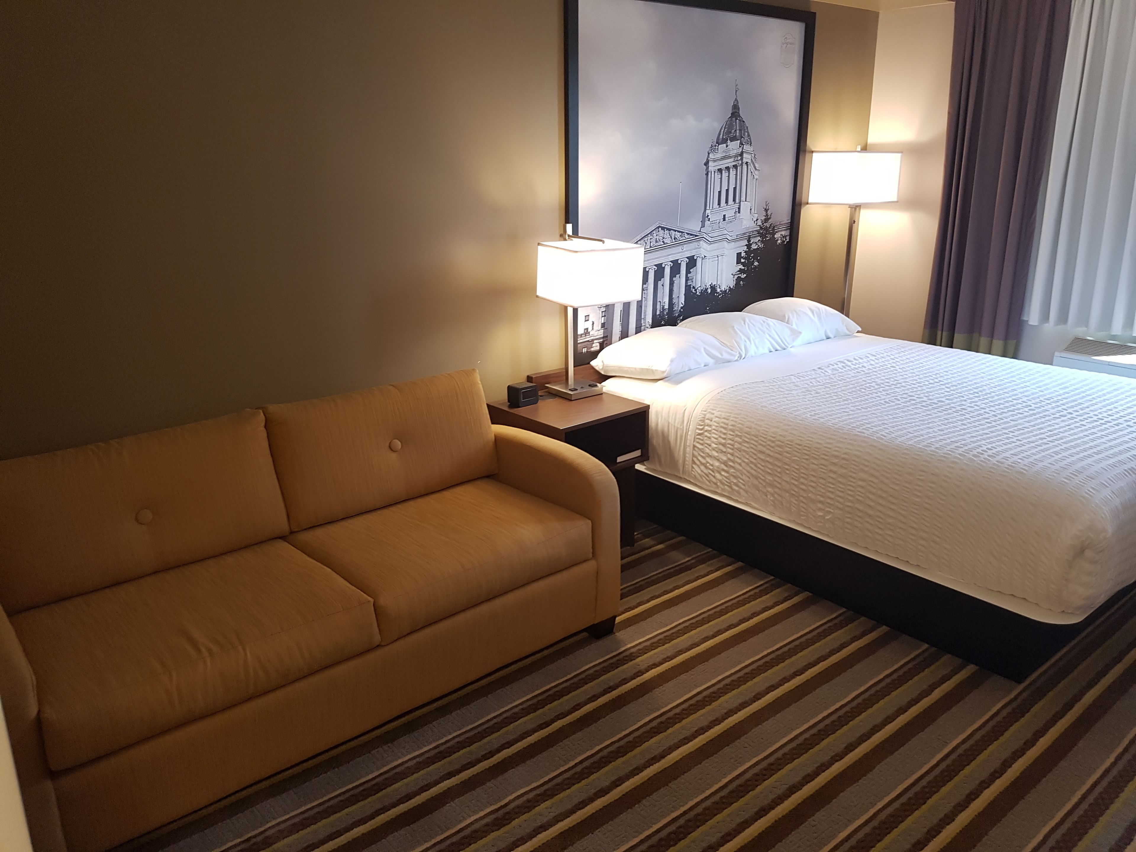A hotel room with a bed, a sofa, a nightstand with a lamp, and a framed picture on the wall.