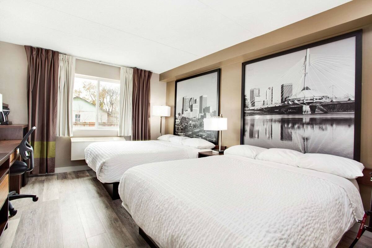 Ein Hotelzimmer mit zwei Betten, einem Schreibtisch, einem Sessel, einem Fenster mit Vorhängen und zwei großen Stadtansichten in Bilderrahmen.