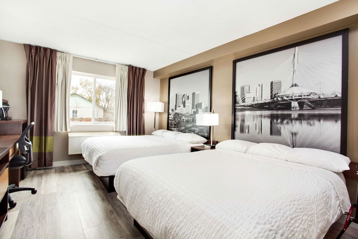 Une chambre d’hôtel avec deux lits, un bureau, une chaise, une fenêtre avec des rideaux et deux grandes photos encadrées représentant des vues de la ville.