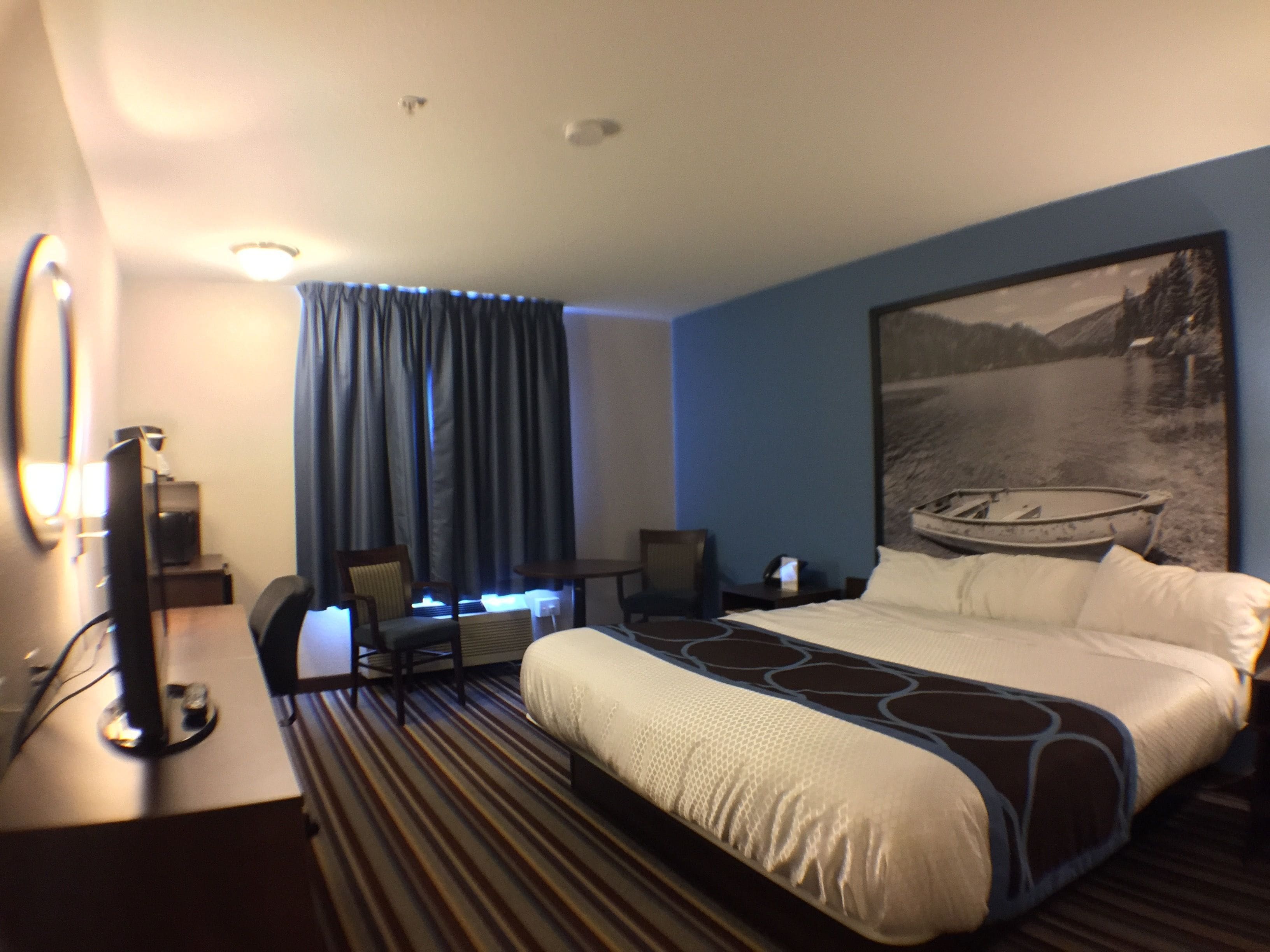 A hotel room with a bed, a desk, a chair, a television, and a large painting of a boat on a lake.