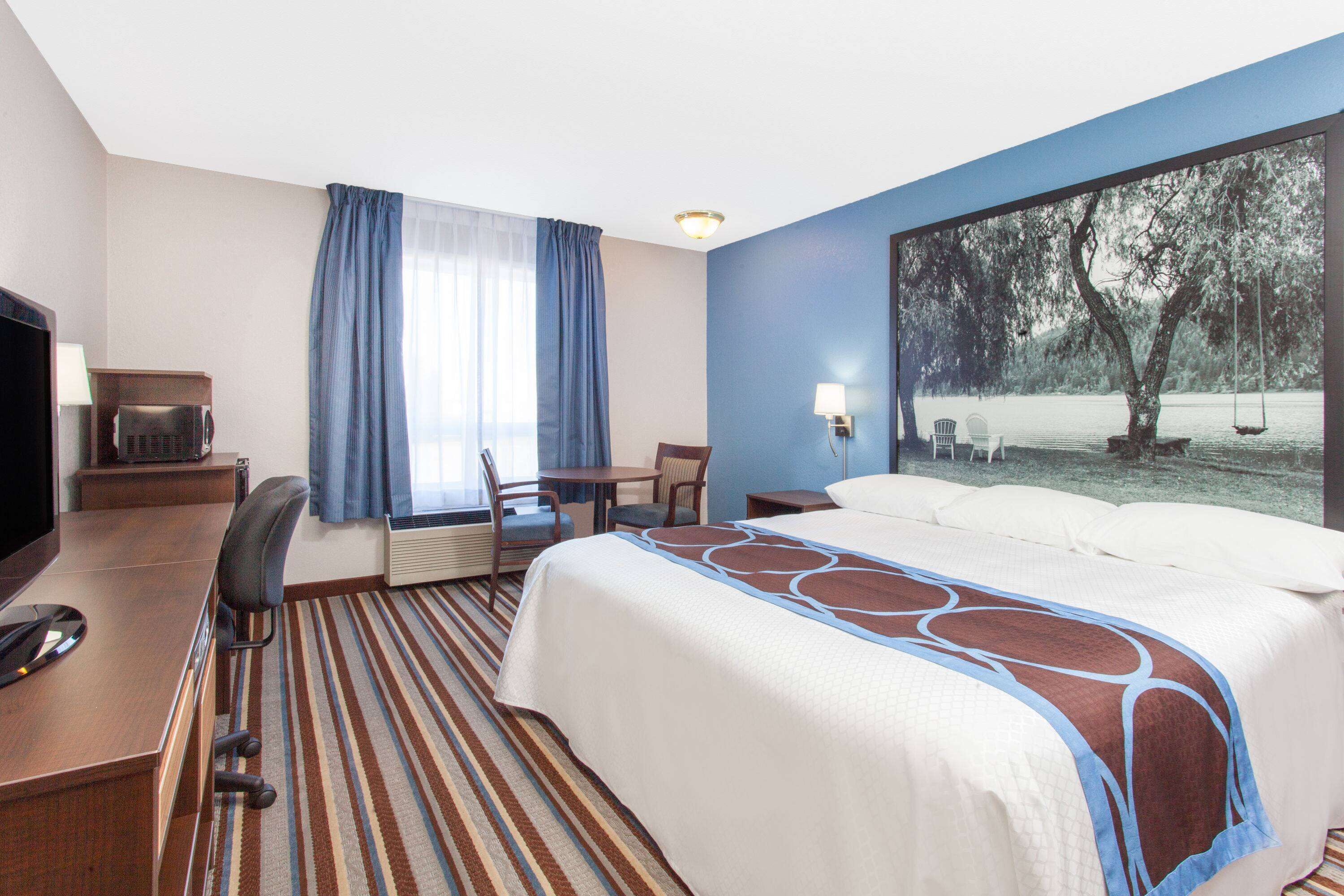 Habitación de hotel con una cama grande, un escritorio con computadora, una silla, un televisor y un ventanal con cortinas.