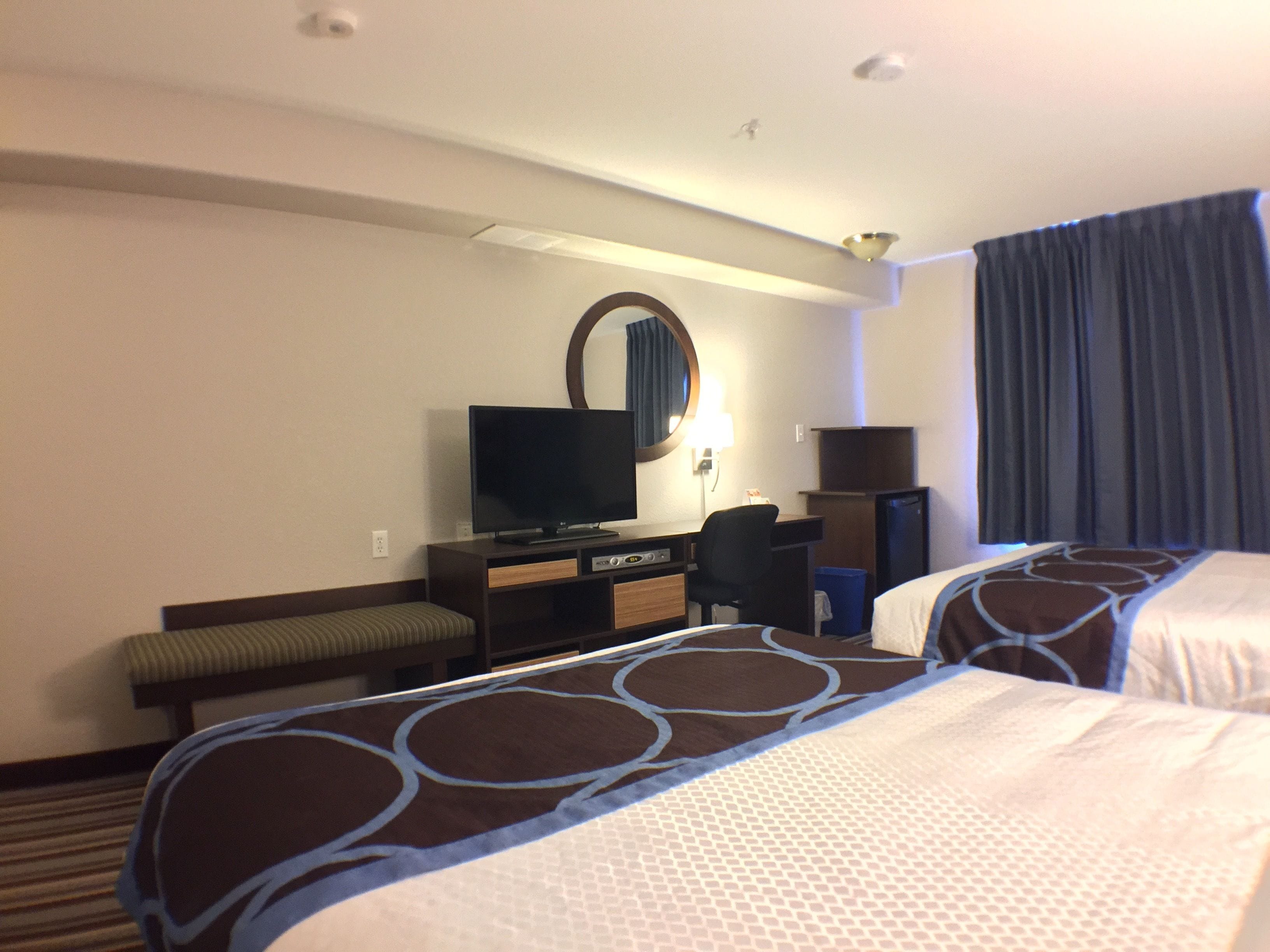 A hotel room with two beds, a television, a desk with a chair, a mirror, and a window with curtains.