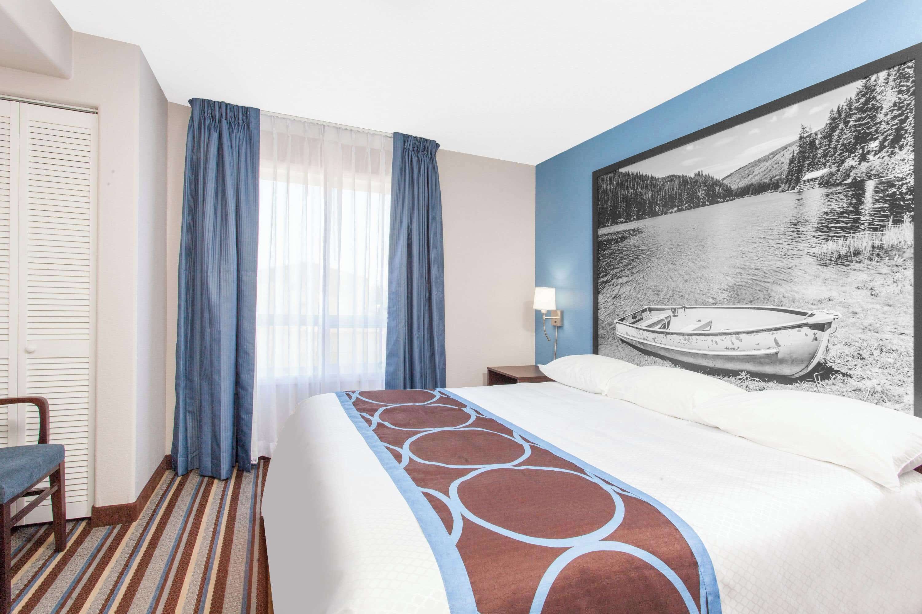 Une chambre d’hôtel avec un lit, une chaise, une fenêtre avec des rideaux et un tableau représentant un bateau sur un lac, accroché au mur.