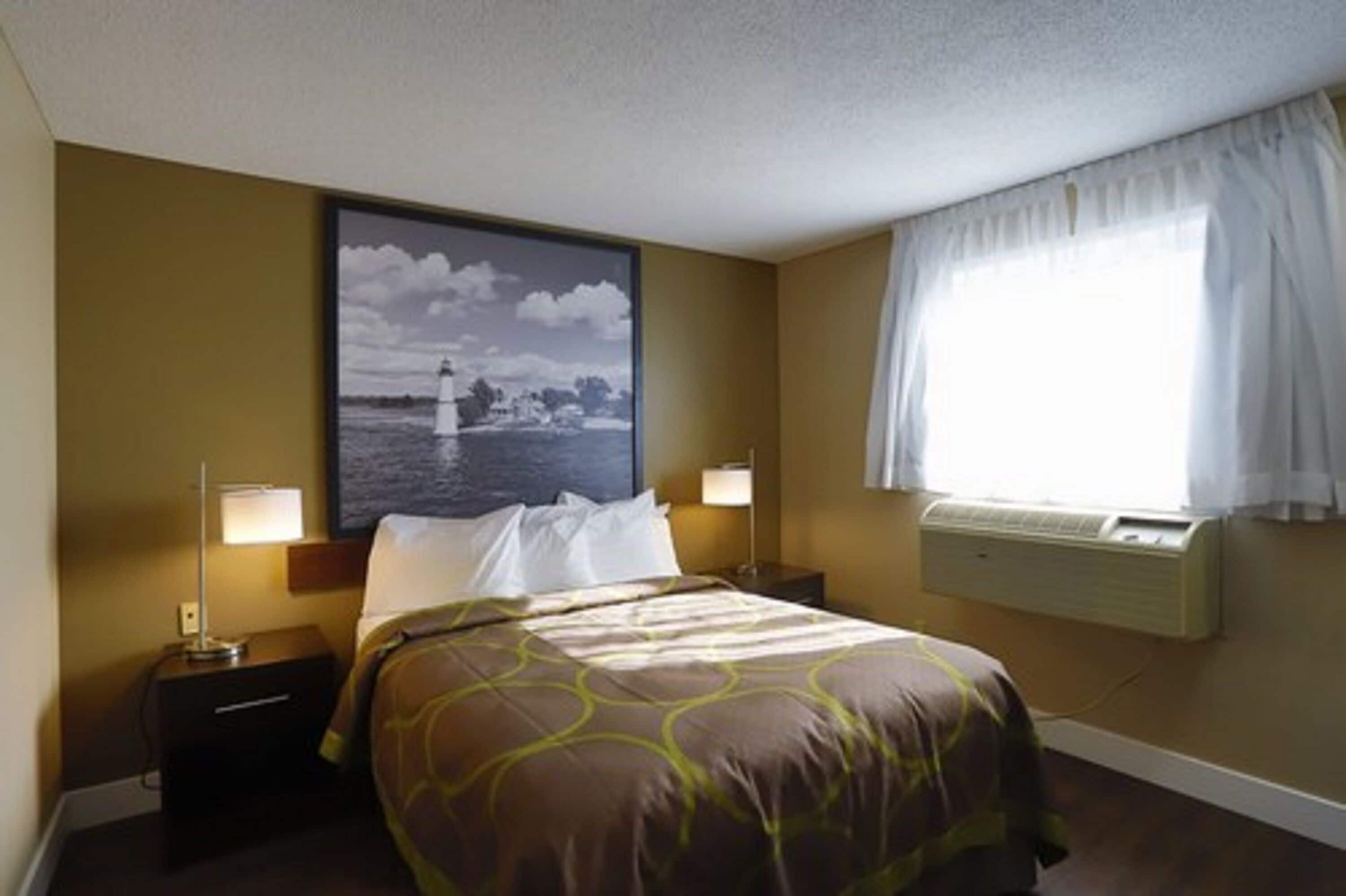 A hotel room with a bed, two bedside lamps, a nightstand, a framed picture on the wall, a window with curtains, and an air conditioning unit.