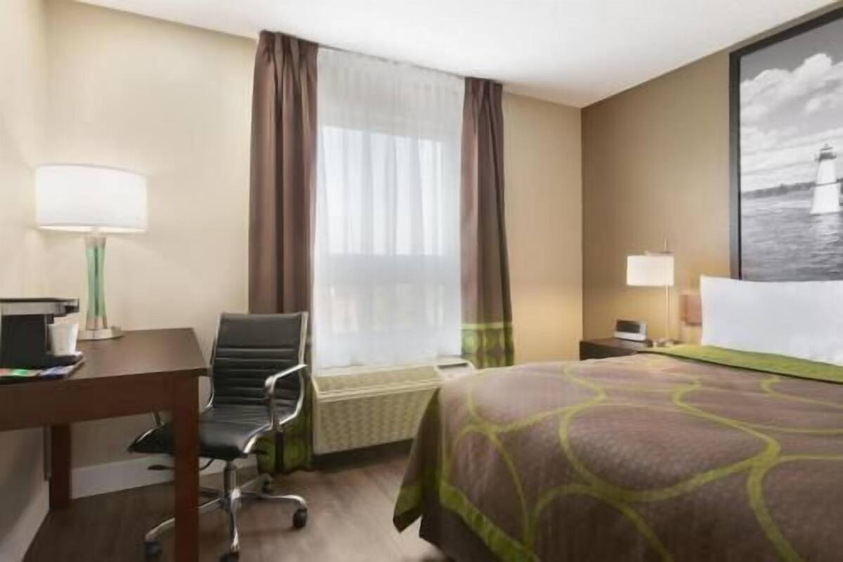 Ein Hotelzimmer mit Bett, Schreibtisch mit Stuhl, Fenster mit Vorhängen, einer Lampe und einem Bild an der Wand.