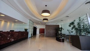 Un hall d’hôtel moderne avec un comptoir d’accueil en marbre, un espace salon avec des fauteuils, une grande plante en pot et un mur orné d’un motif géométrique.