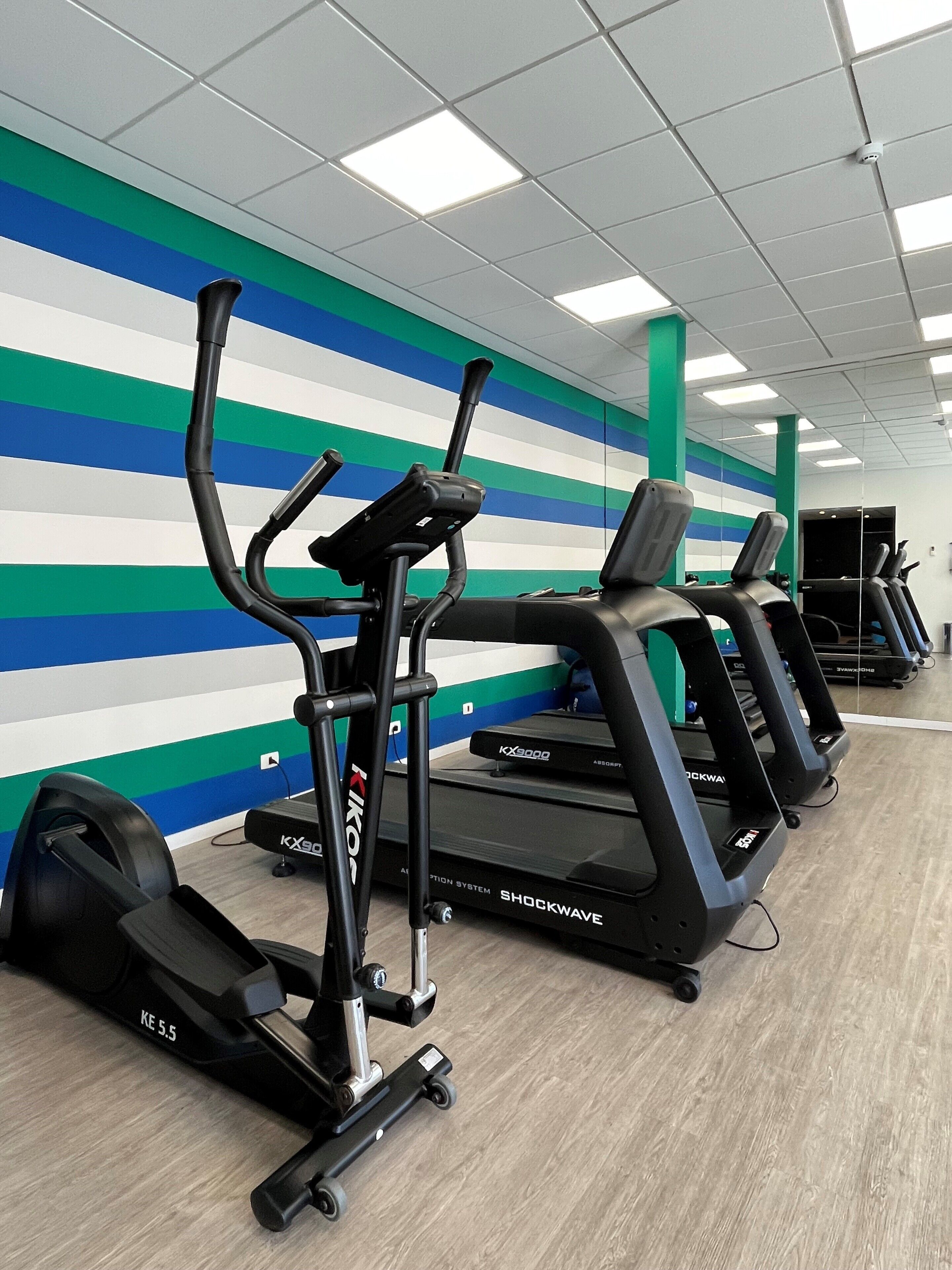 En rad löpband och crosstrainers i ett gym med en randig vägg och takbelysning.