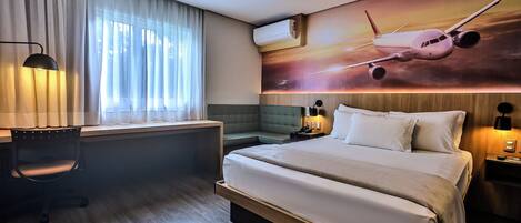 A hotel room with a large bed, a desk with a chair, a window with curtains, an air conditioning unit, a nightstand with a lamp, and a wall-mounted painting of an airplane.