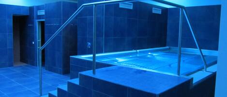 A blue-lit indoor pool area with a glass enclosure, steps leading up to it, and a door in the background.