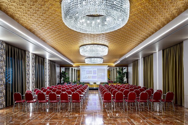 Un salón de conferencias con filas de sillas rojas, una gran lámpara de araña y una pantalla de presentación al frente.