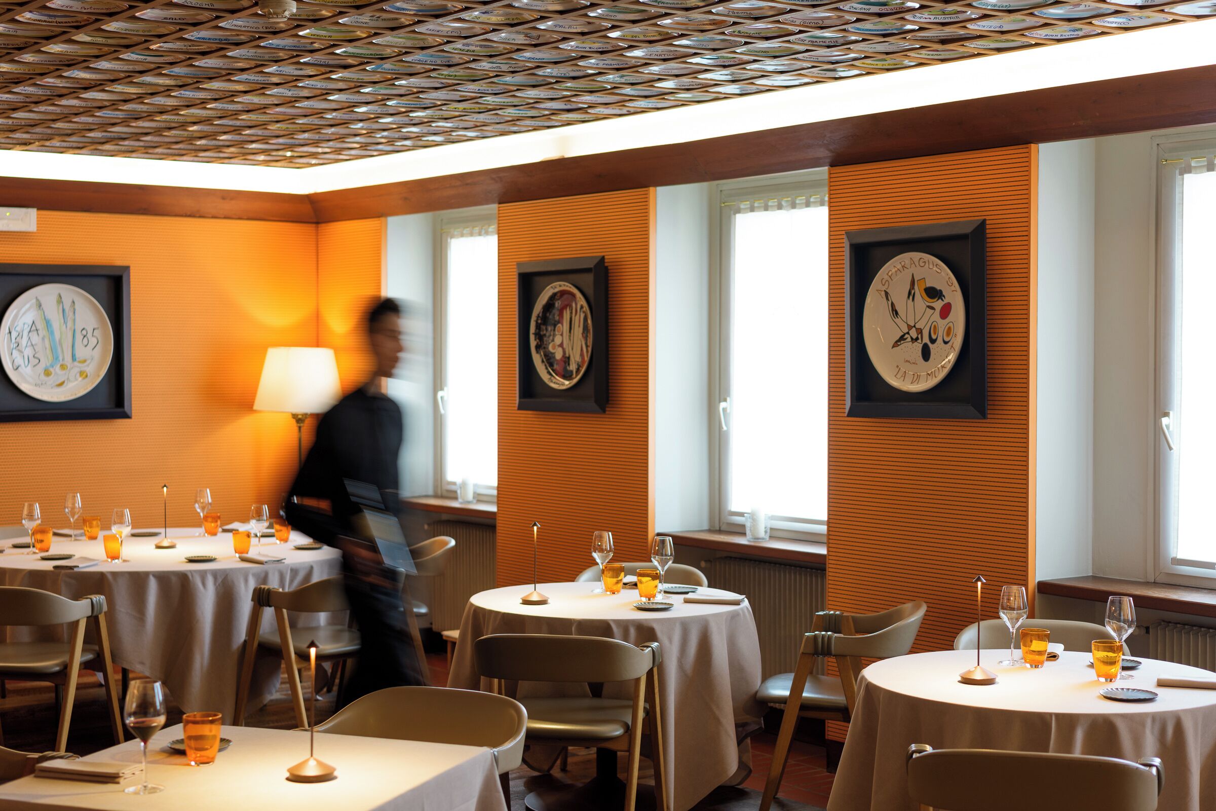A restaurant with round tables, framed artwork on the walls, and a patterned ceiling.