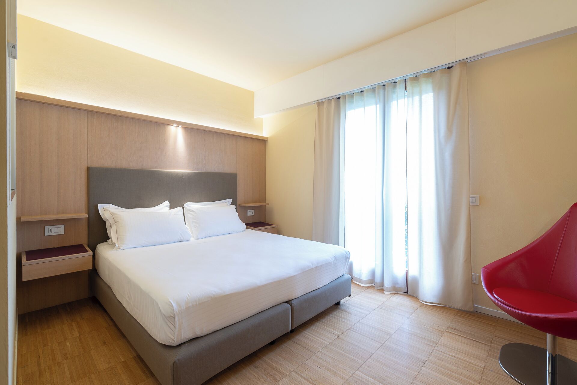 Quarto de hotel moderno com uma cama grande, painéis de madeira, uma cadeira vermelha e vista para o exterior.