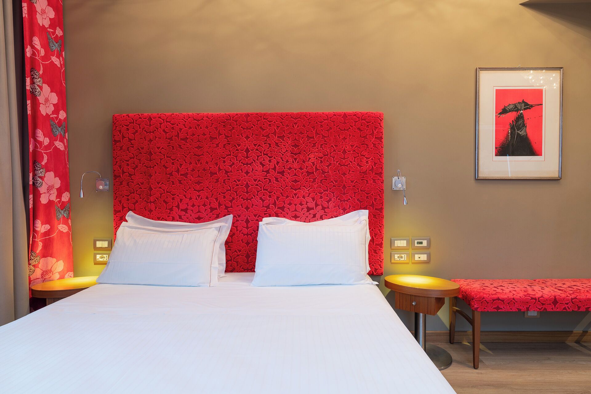 A hotel room with a large bed, two bedside tables, a bench, a wall-mounted artwork, and a red floral curtain.