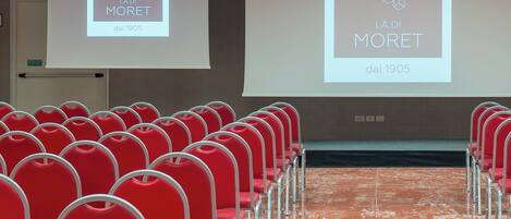 A conference room with rows of red chairs and a large screen displaying the logo "Là Di Moret dal 1905".