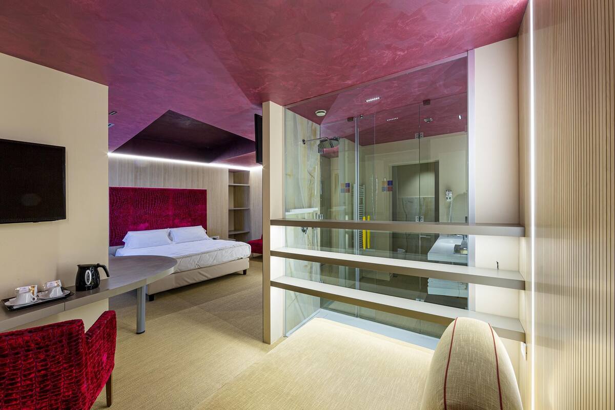 Une chambre d’hôtel moderne avec une cloison en verre, un lit, un bureau avec une chaise et un fauteuil rouge recouvert de tissu.