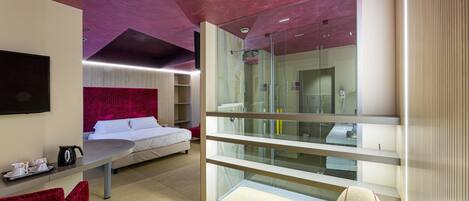 A modern hotel room with a glass partition, a bed, a desk with a chair, and a red upholstered chair.