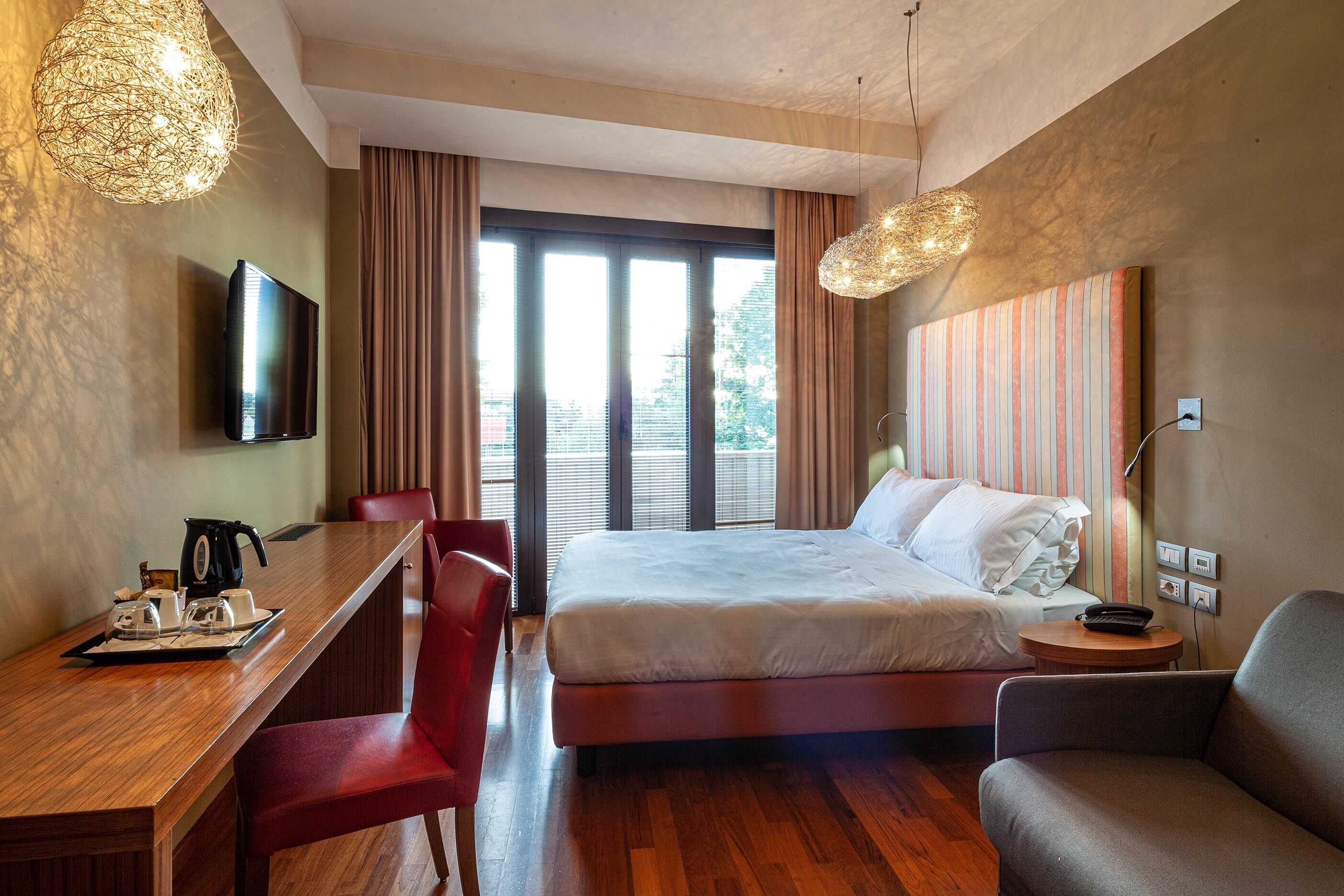 A modern hotel room with a wooden desk, a bed, a TV, and a large window with curtains.