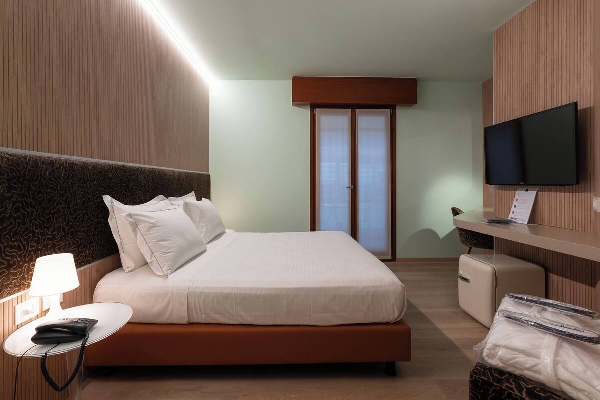 Une chambre d’hôtel avec un lit, une table de chevet avec une lampe, un téléviseur fixé au mur et une valise posée sur le sol.