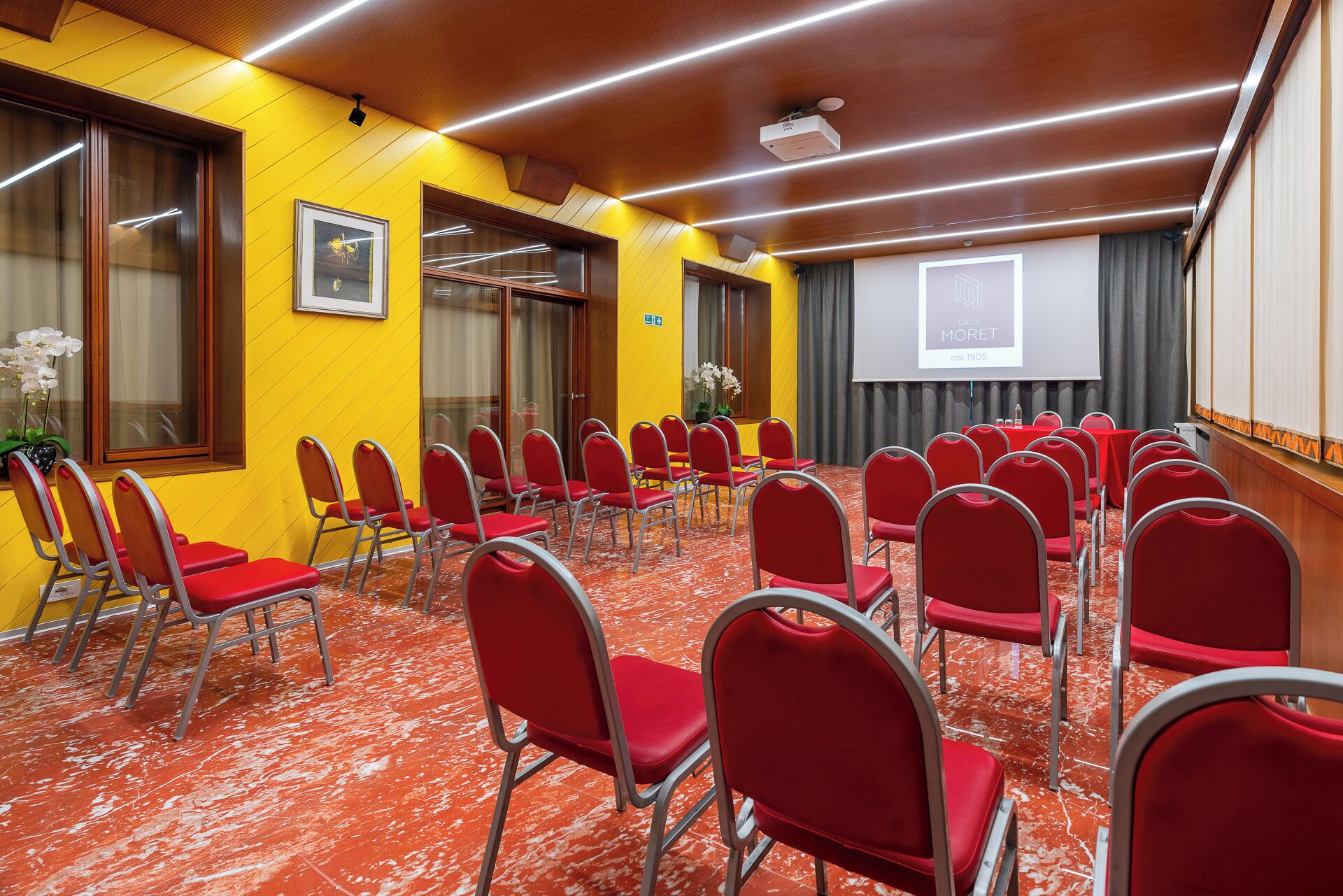 Un salón de conferencias con filas de sillas rojas, una pantalla de proyector y una pared amarilla.