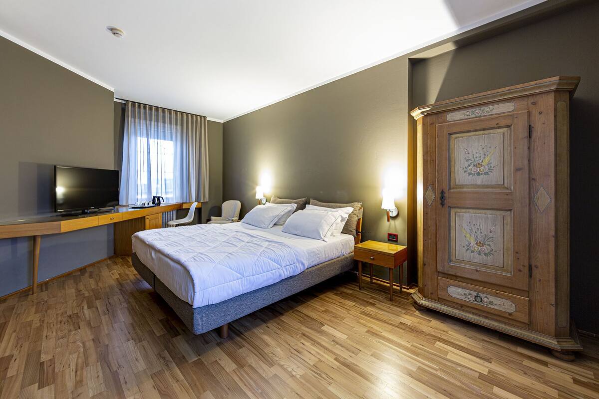 Une chambre à coucher comprenant un lit, un bureau avec une télévision, une armoire et une fenêtre avec des rideaux.
