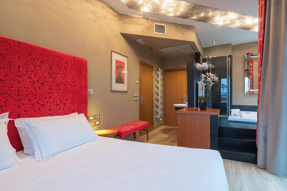 Une chambre d’hôtel avec un grand lit, un mur d’accent rouge, une petite table, un banc, une salle de bain avec un miroir et un espace douche.