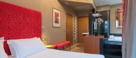 A hotel room with a large bed, a red accent wall, a small table, a bench, a bathroom with a mirror, and a shower area.