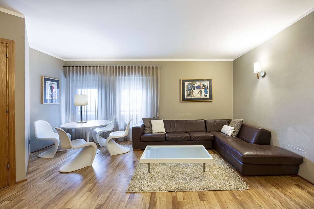 Un salon moderne avec un canapé en cuir marron, une table basse blanche et une table à manger ronde entourée de chaises blanches. Deux œuvres d’art encadrées ornent les murs, et une lampe se trouve sur le côté droit.