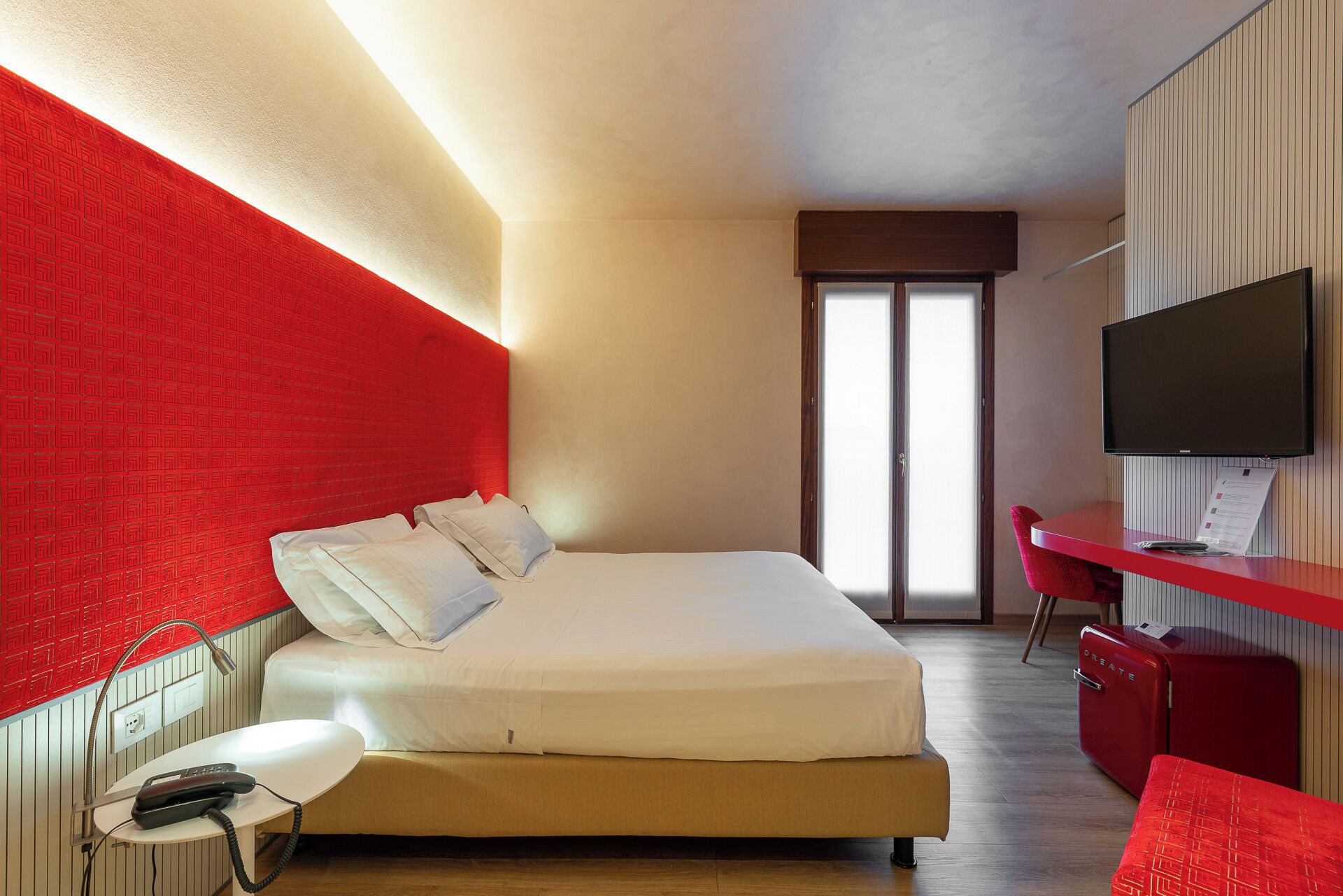 A hotel room with a large bed, a desk with a chair, a television, and a red suitcase.