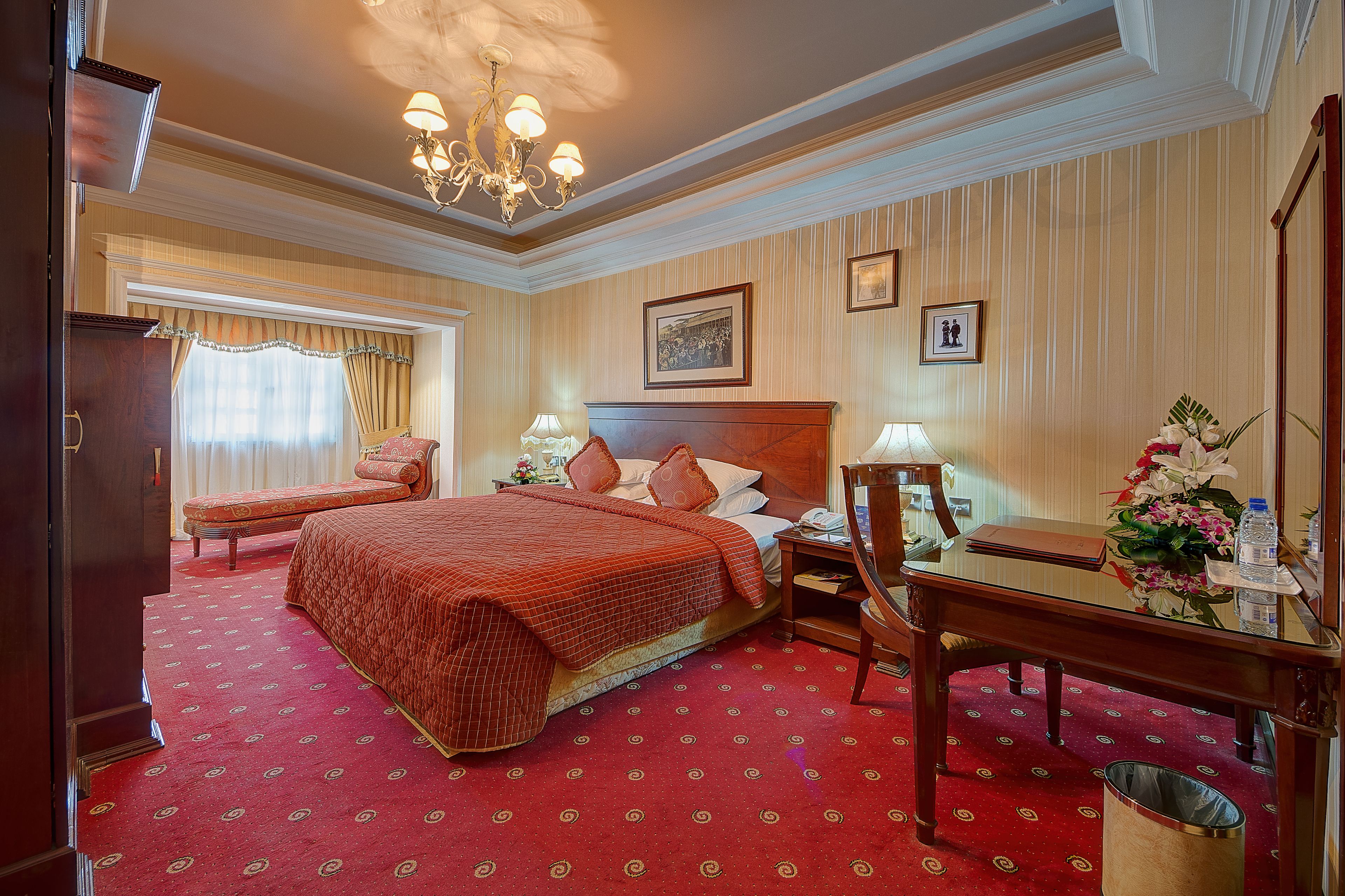 A hotel room with a large bed, a desk, a chair, a nightstand, a lamp, and a trash can.
