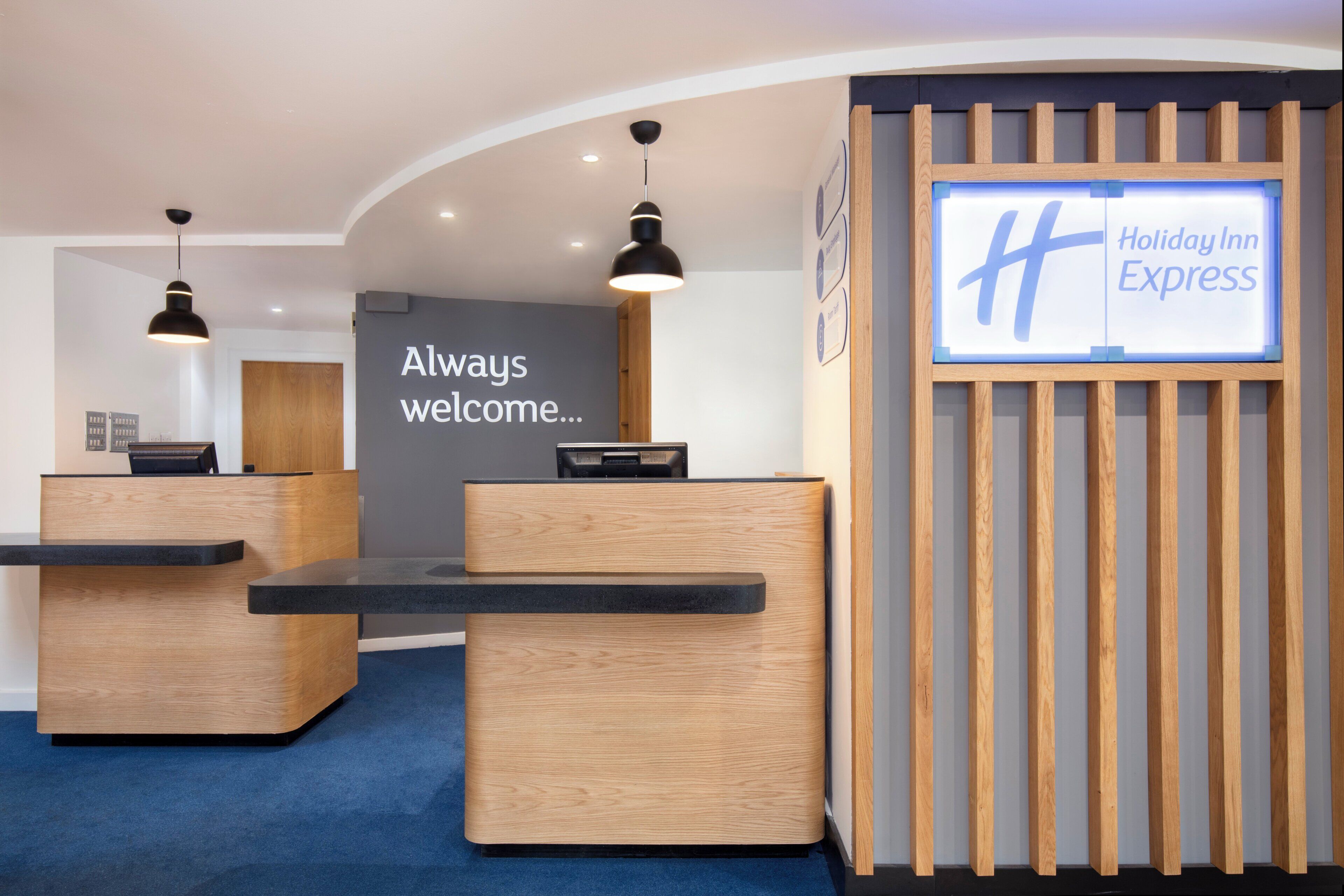 A reception area with a wooden reception desk, a digital display board, and a wall with a welcoming message.