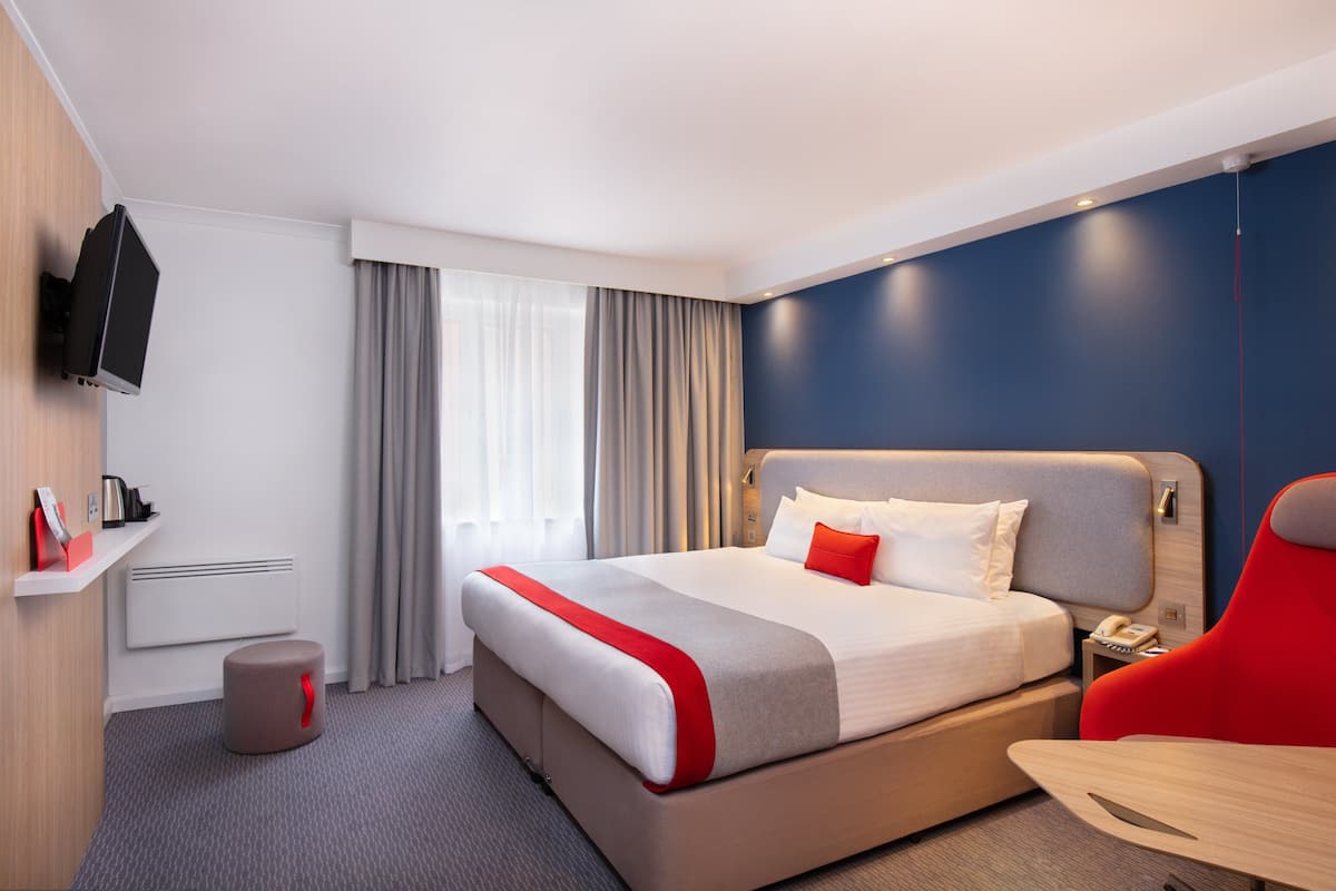 A modern hotel room with a large bed, a red chair, a flat-screen TV mounted on the wall, and a small table.