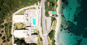 An aerial view of a coastal property with a swimming pool, a large building with a unique roofline, and a beach area. - Canvas by Mitsis Belvedere (Corfu)