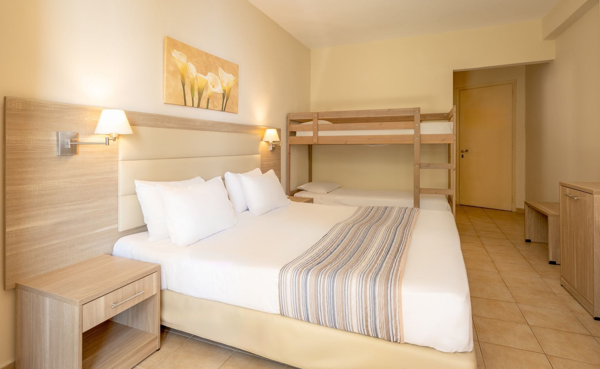 A hotel room with a bunk bed, a bedside table with a lamp, a painting on the wall, and a door.