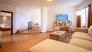 A modern living room with a sofa, a coffee table, a TV mounted on the wall, and a dining area with a table and chairs.